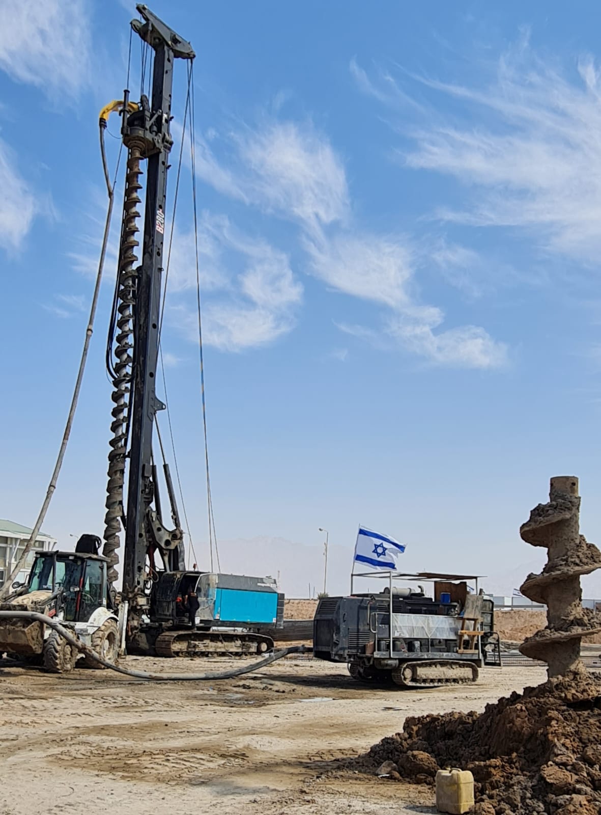 מכונת קידוח וספריח באתר בנייה במדבר עם דגל ישראל והרי אילת ברקע