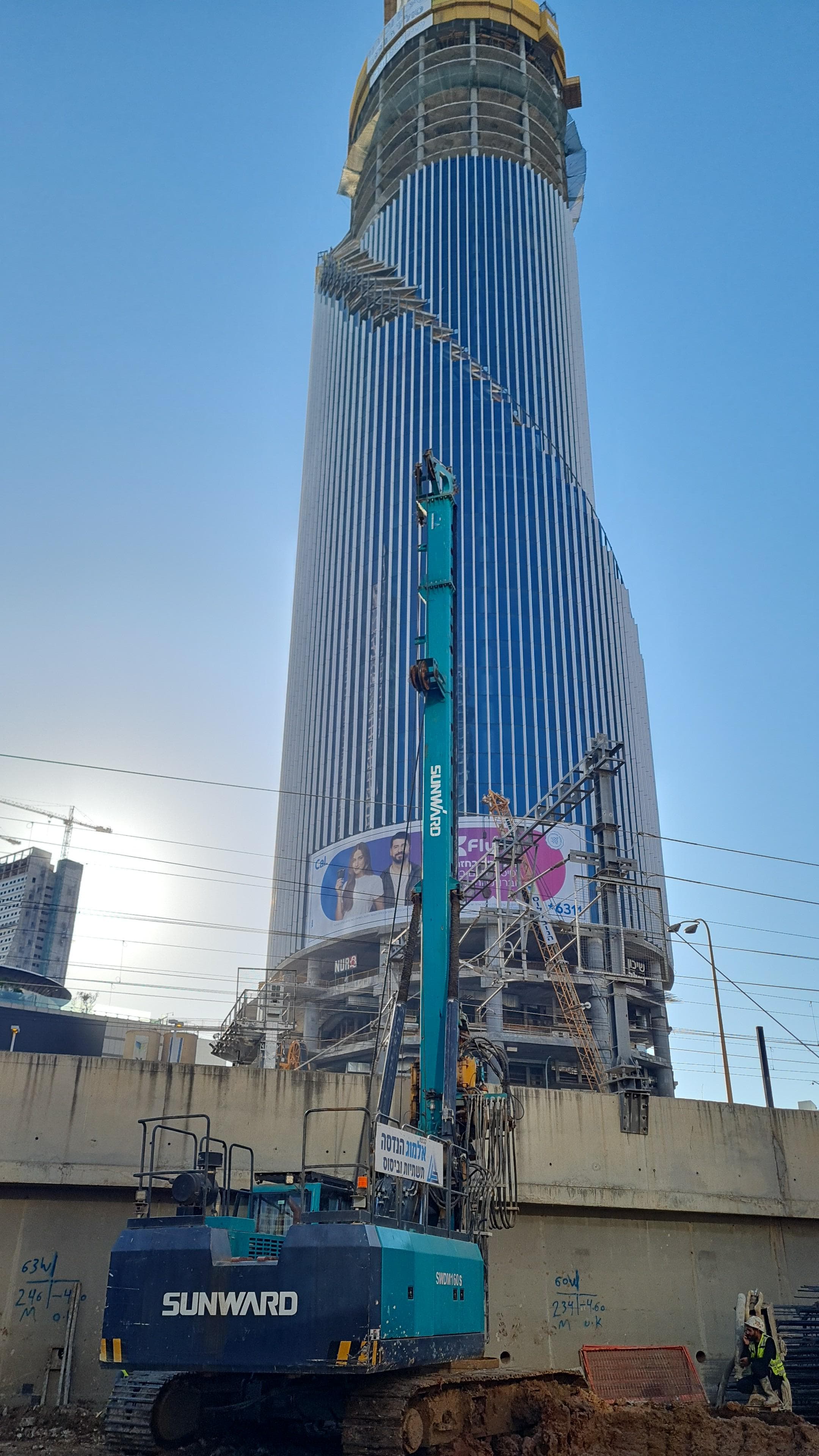 מכונת קידוח ליד מגדל רב-קומות בתל אביב, עם שילוט ופיגומי בנייה ברקע