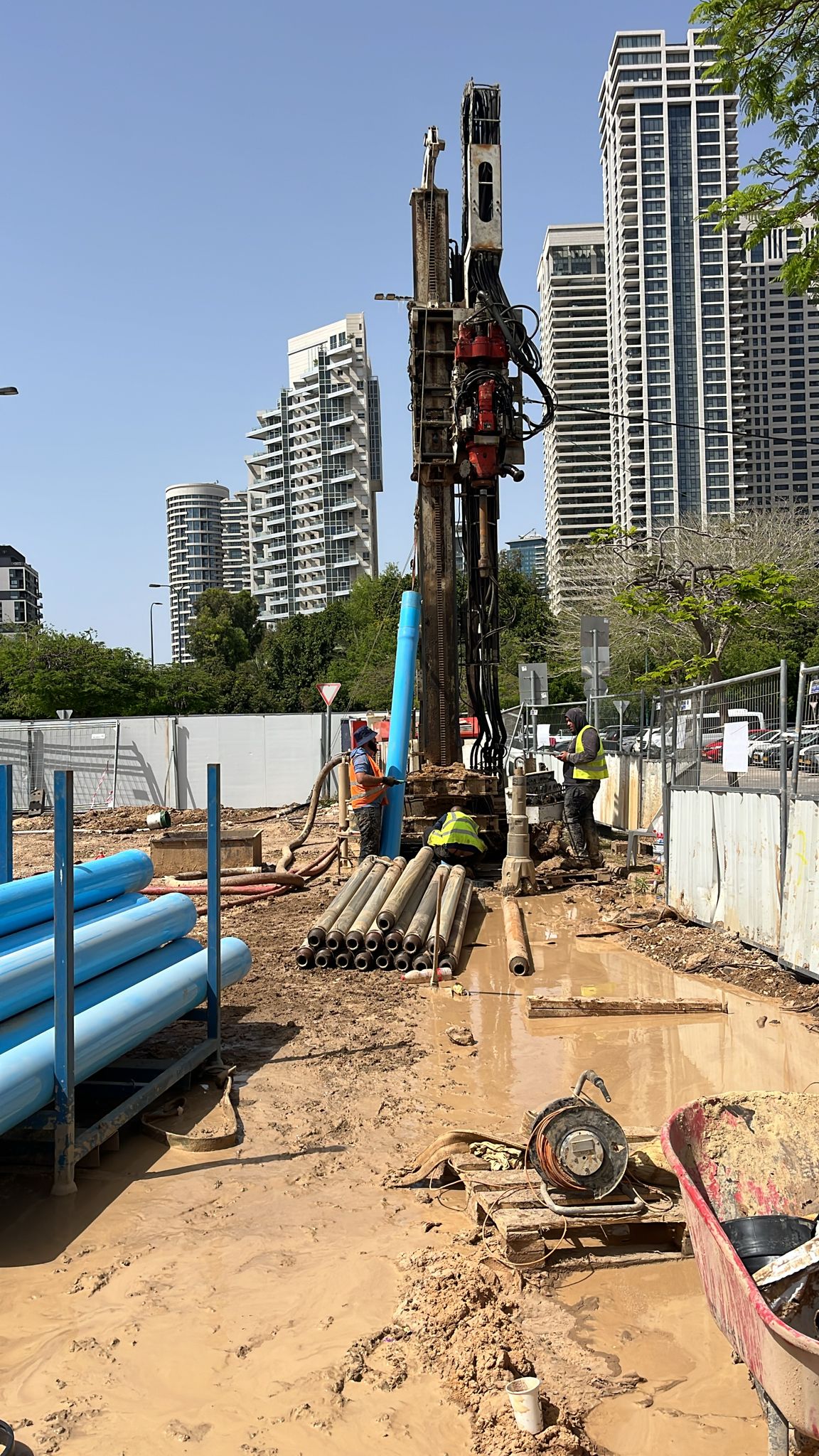 מכונת קידוח מתקינה צינור פיאזומטר כחול לצד רחוב עירוני בתל אביב, עם מגדלי משרדים ברקע