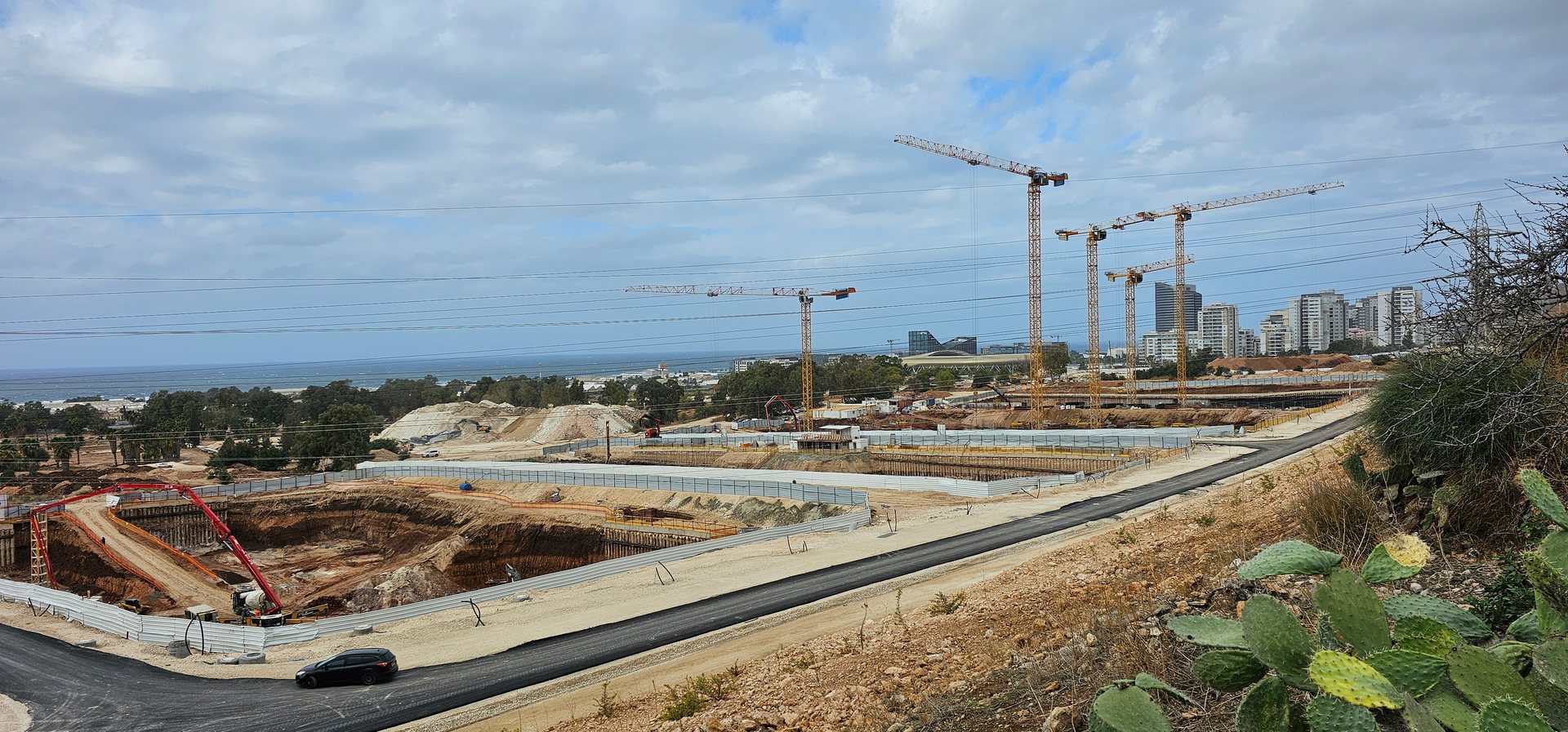 אתר בנייה נרחב עם מספר עגורני מגדל וחפירות ביסוס, ברקע קו החוף וקו הרקיע של חיפה
