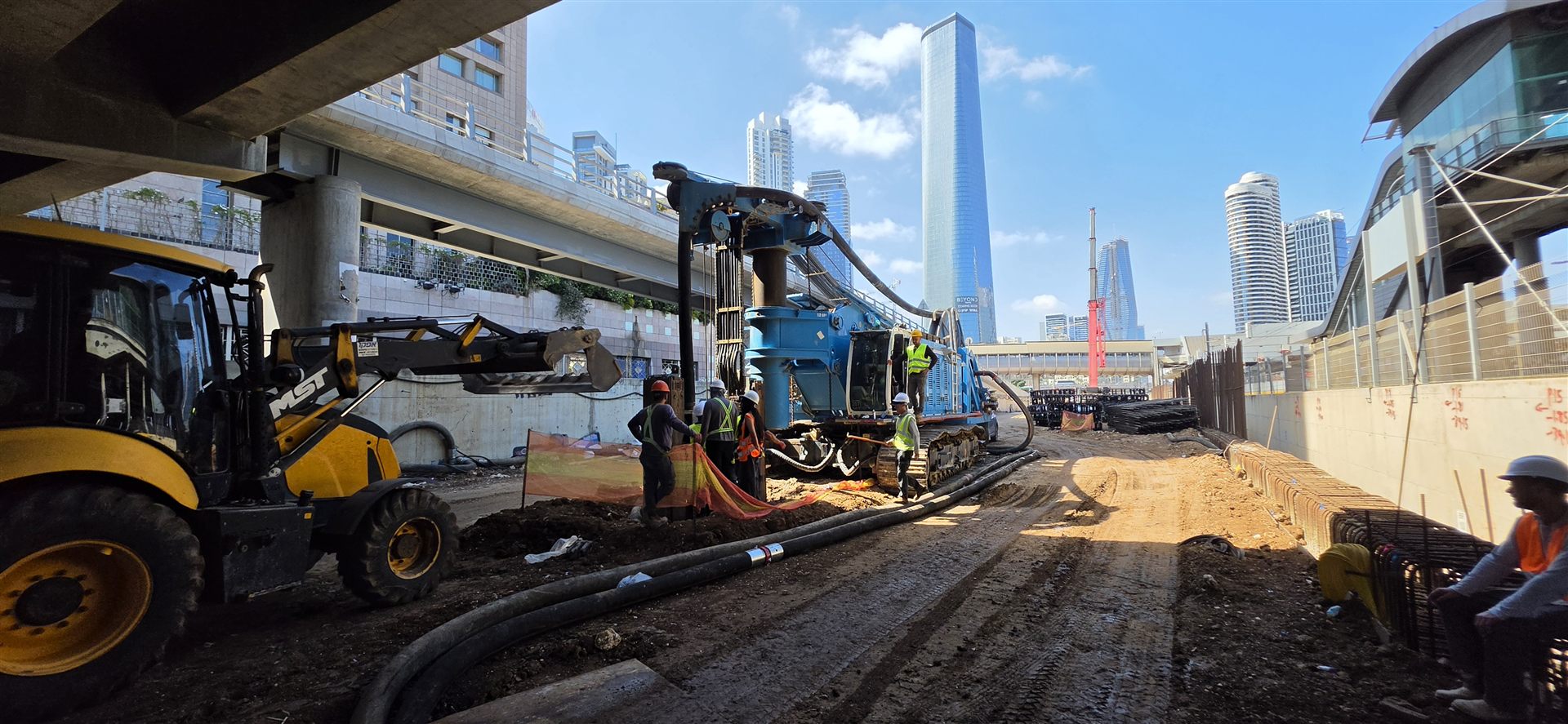 מכונת קידוח סיבובית פועלת מתחת לגשר תחבורה בלב תל אביב, עם מגדל עזריאלי שרד וקו הרקיע של העיר ברקע