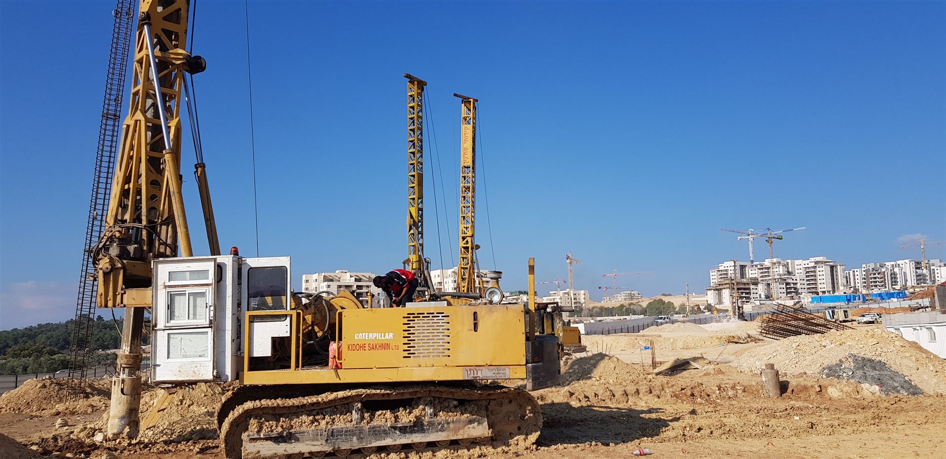 מכונת קידוח סיבובית עם שני תרנים באתר ביסוס, ברקע בנייני מגורים בשלבי הקמה ועגורני מגדל