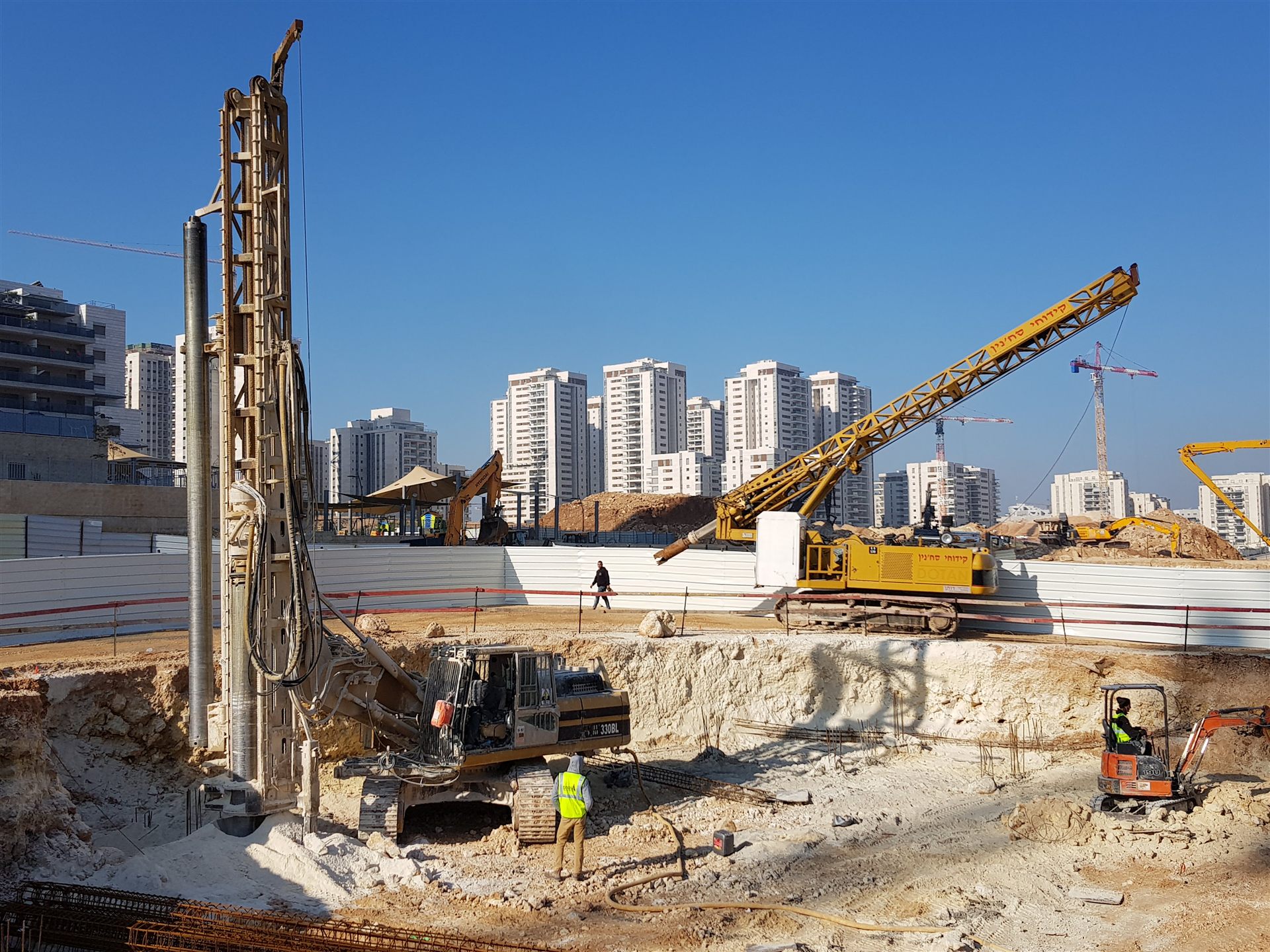 מכונת קידוח סיבובית צהובה באתר ביסוס, ברקע בנייני מגורים גבוהים וקו רקיע עירוני