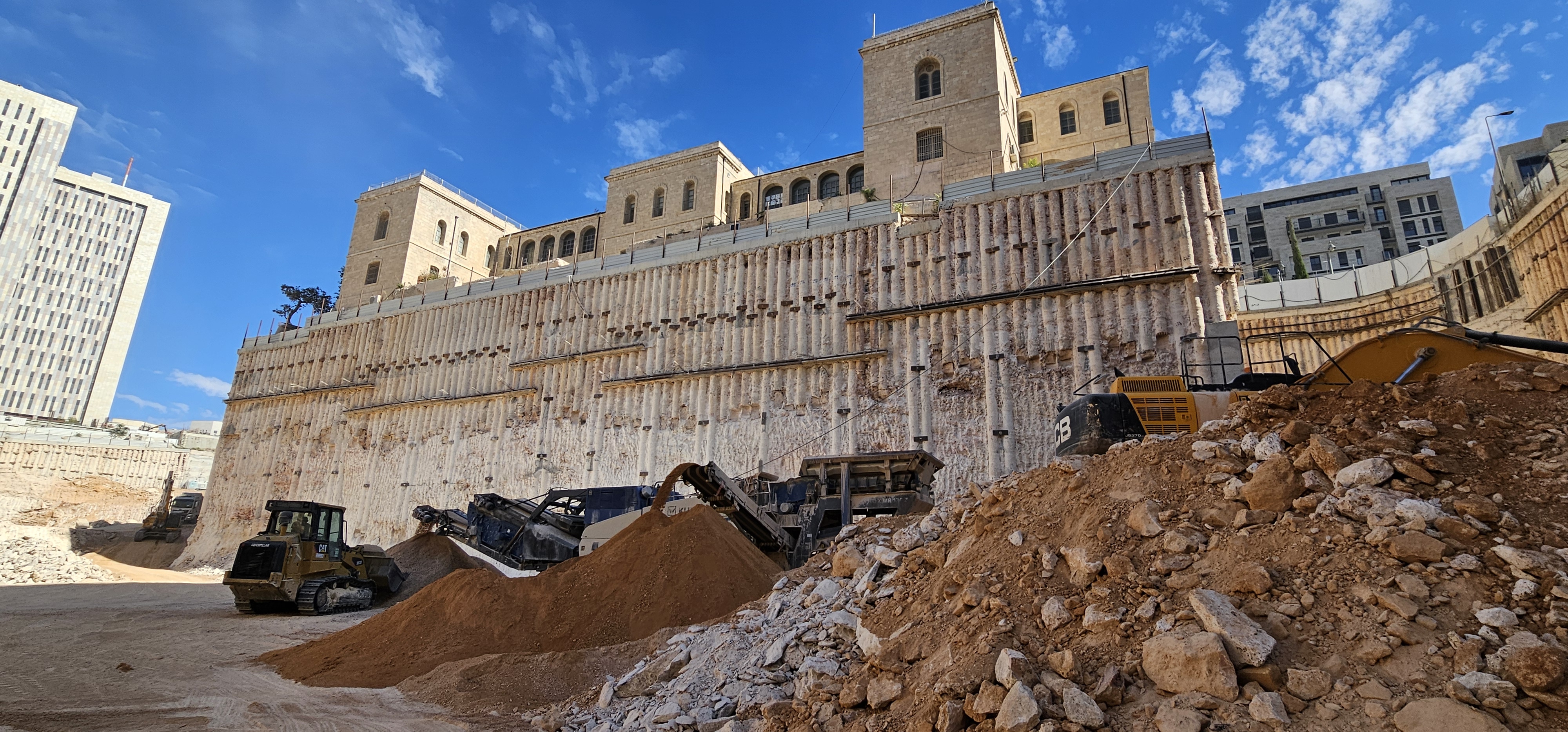 קיר דיפון מקידוחים תומך בחפירה עמוקה מתחת למבנה אבן היסטורי, עם ציוד כרסום בחזית