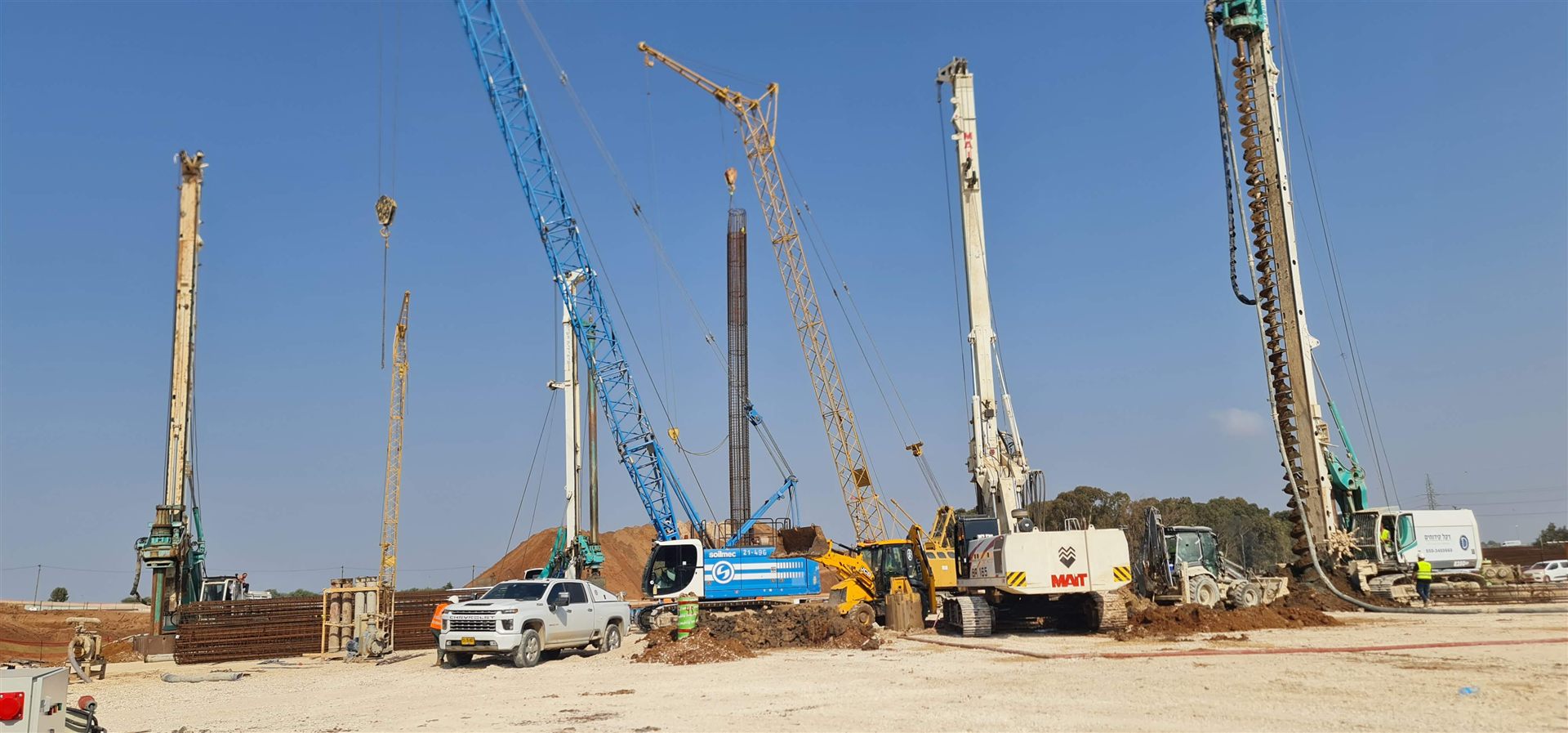 צוות עובדים ומכונות קידוח באתר ביסוס נרחב, עם כלובי זיון וכלי רכב בשטח