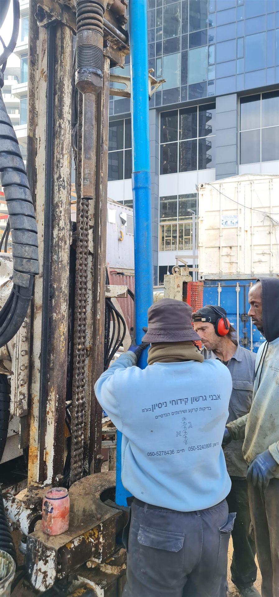 עובד מתקין צינור פיזומטר כחול במכונת קידוח באתר עירוני, עם מגדלים גבוהים ברקע