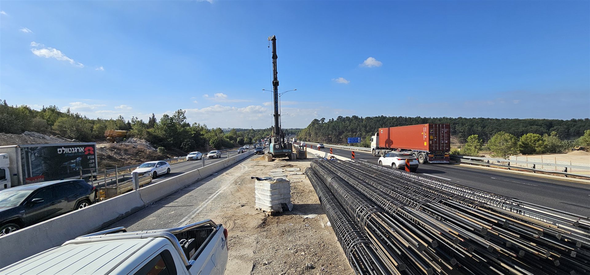 עבודות הקמה על גשר כביש 6, עם כלובי זיון, משאיות ומכונת קידוח בצד הכביש