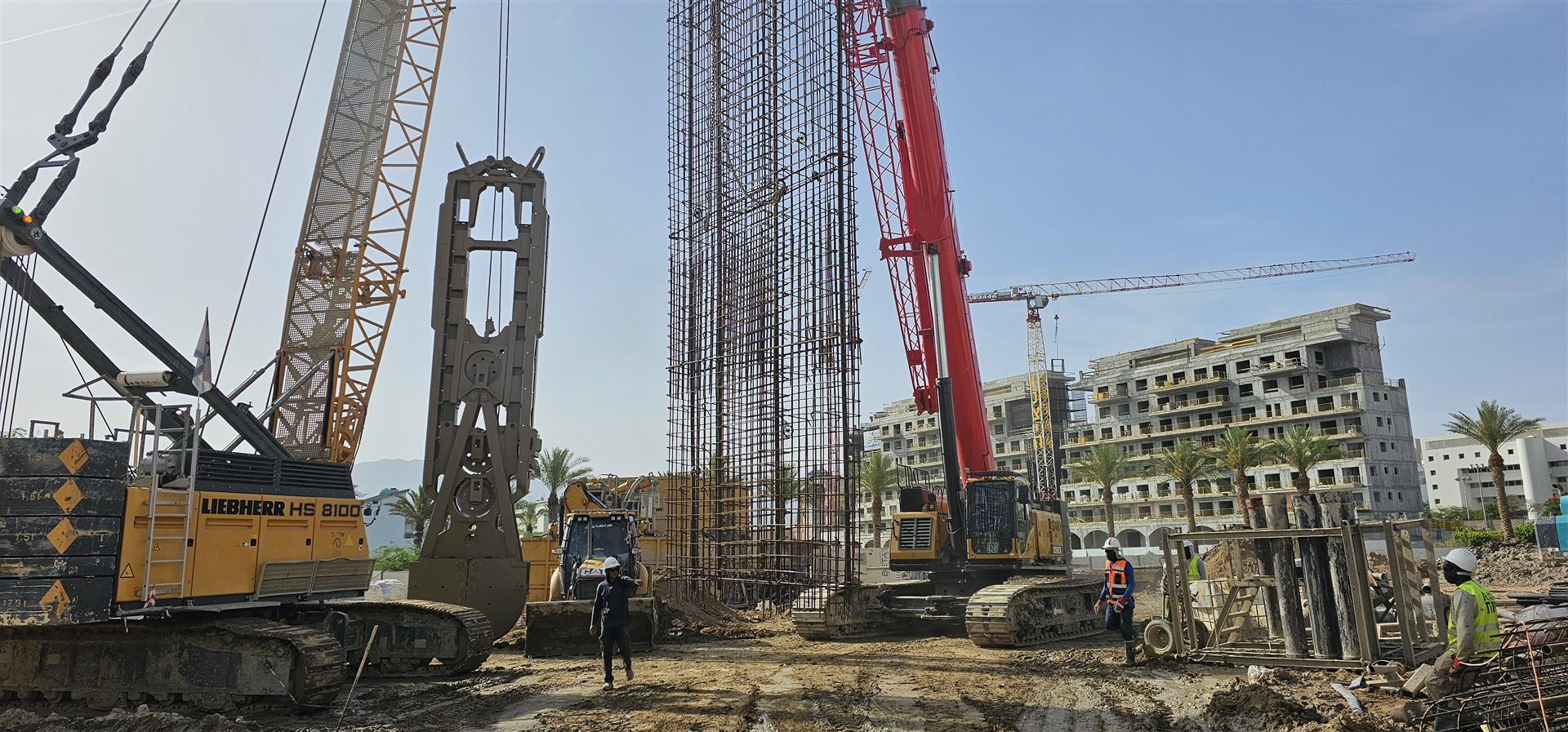 אתר בנייה עם עגורנים וכלוב זיון, דקלים ובנייני מגורים בשלבי הקמה ברקע