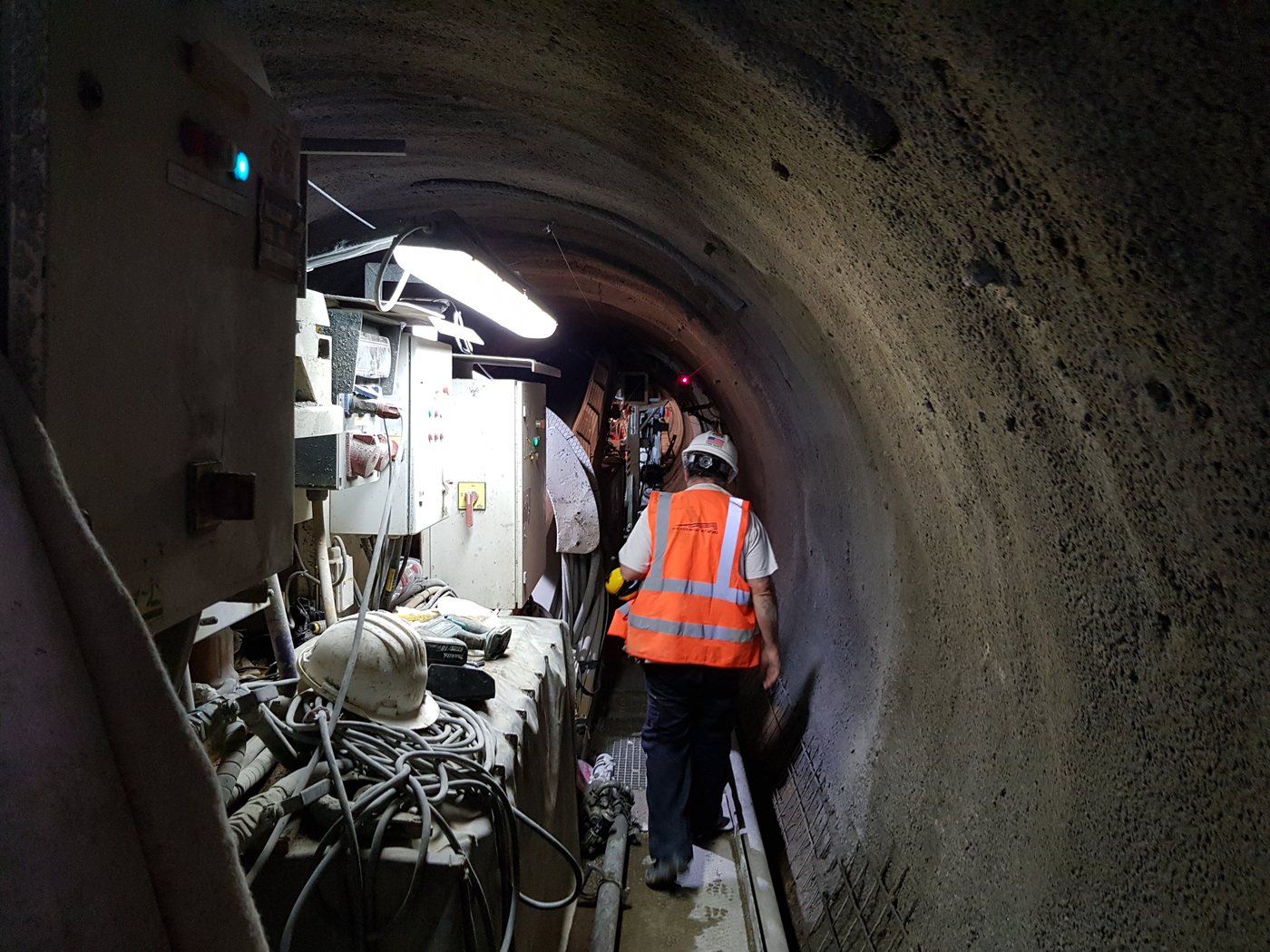 עובד עם אפוד זוהר הולך במנהרת TBM מוארת, עם ציוד הכרייה וארון חשמל בחזית